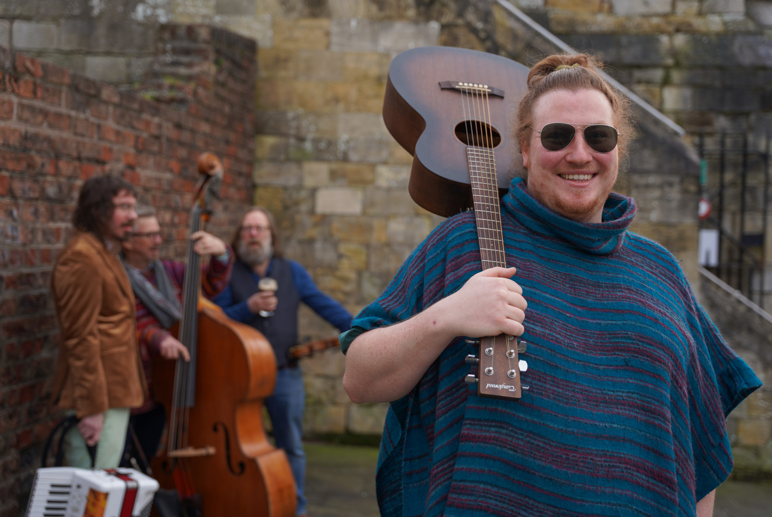 Man in front wearing a blue jumper and sunglasses, smiling and holding an acoustic guitar over his shoulder. Three men in the background, all holding various instruments.