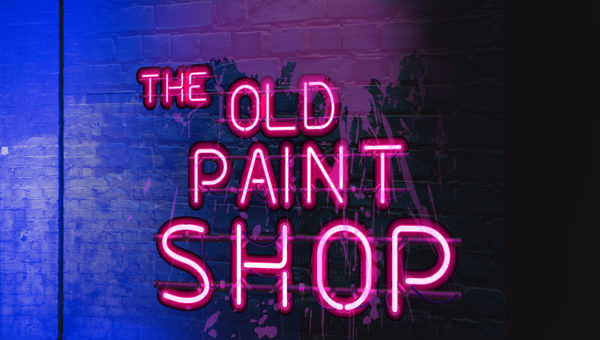 'The Old Paint Shop' in pink neon letters against a dark spray painted wall, with lights hanging above