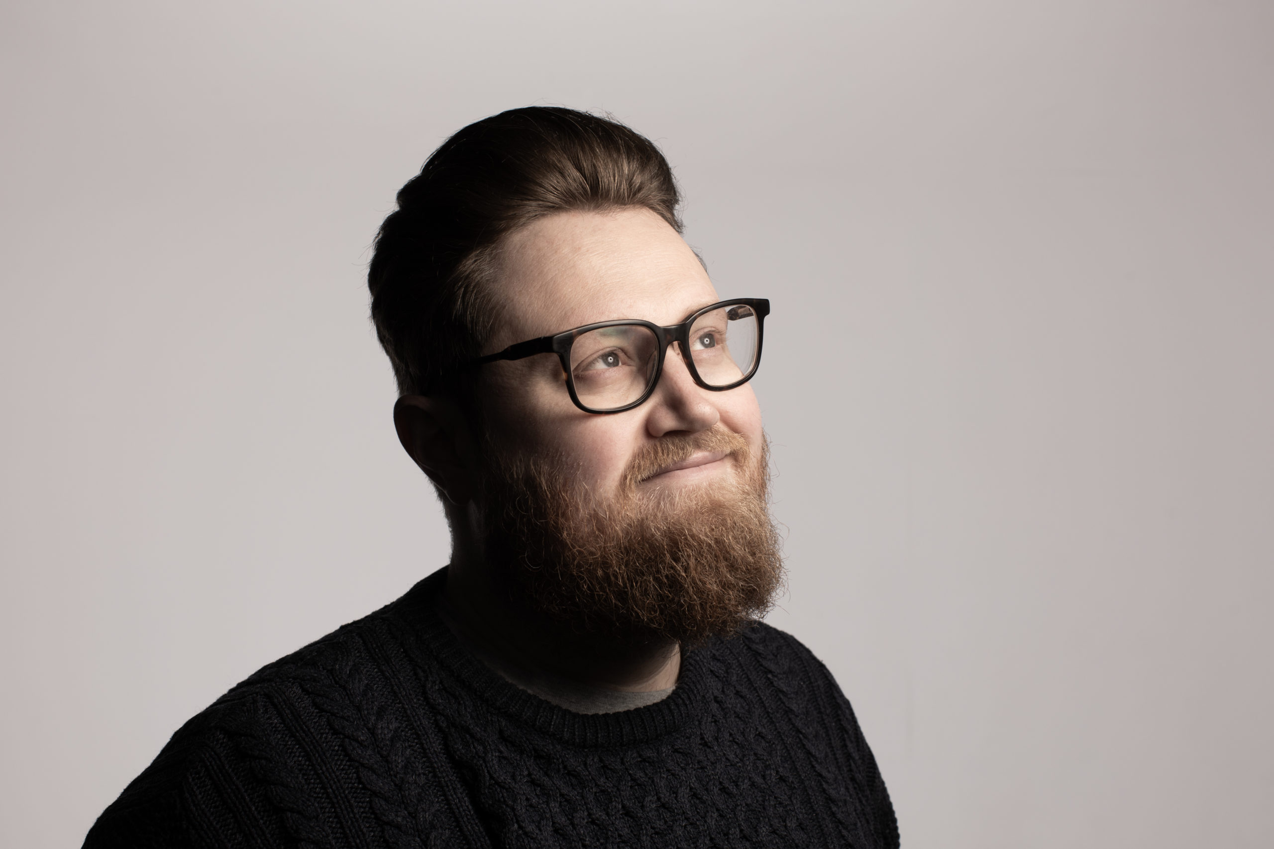Man wearing a black jumper and glasses, stood against plain background, looking off to the right.