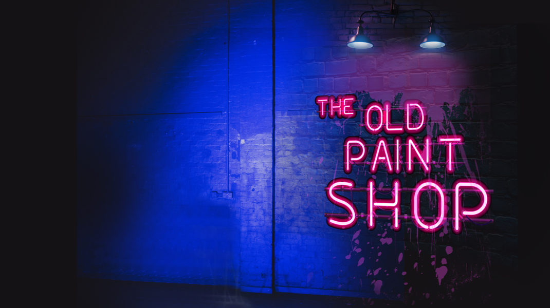 'The Old Paint Shop' in pink neon letters against a dark spray painted wall, with lights hanging above