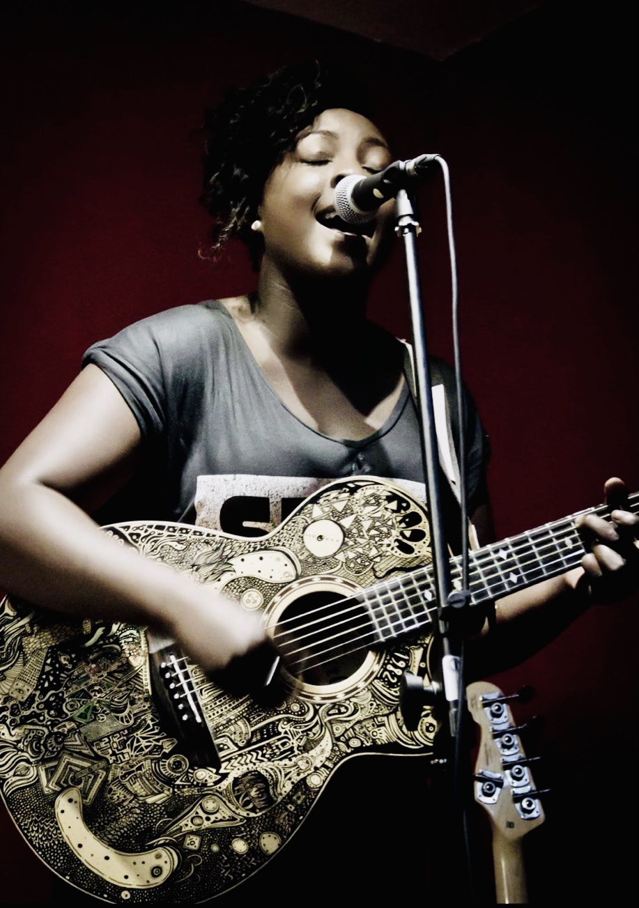 Woman in grey t-shirt singing into a microphone on a stand whilst playing the acoustic guitar.